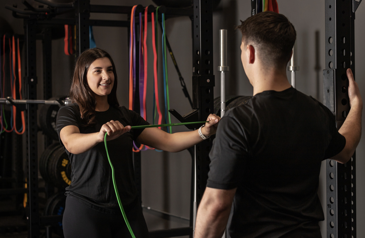 Female physiotherapist instructs male client using green resistance band in modern clinic with colourful equipment storage.